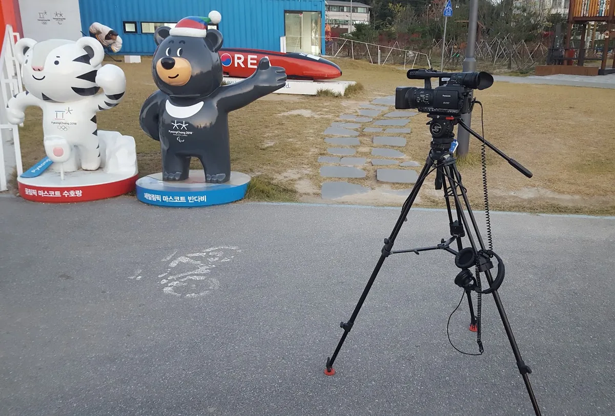 A broadcast camera on a tripod in a park with mascot statues